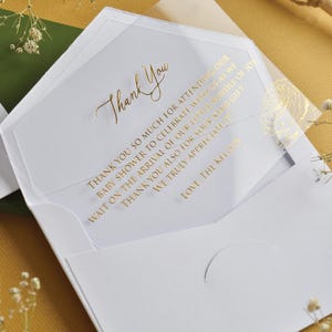 Puede incluir: Una tarjeta de agradecimiento de baby shower blanca con letras doradas dentro de un sobre blanco. La tarjeta dice "Gracias" y un mensaje de gratitud. El fondo es amarillo mostaza.