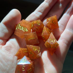 May include: A set of seven orange and gold dice with glitter embedded in the resin. The dice are in various shapes and sizes, including a d4, d6, d8, d10, d12, and d20.
