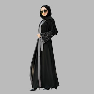 Peut inclure: Une abaya noire avec une bordure grise et des détails en dentelle sur les manches. L'abaya est portée par une personne portant un foulard noir et des lunettes de soleil.