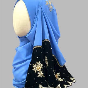 Puede incluir: Un hiyab azul claro con ribete de terciopelo negro y adornos dorados y de perlas. El hiyab tiene un borde festoneado y est&aacute; decorado con dise&ntilde;os florales.
