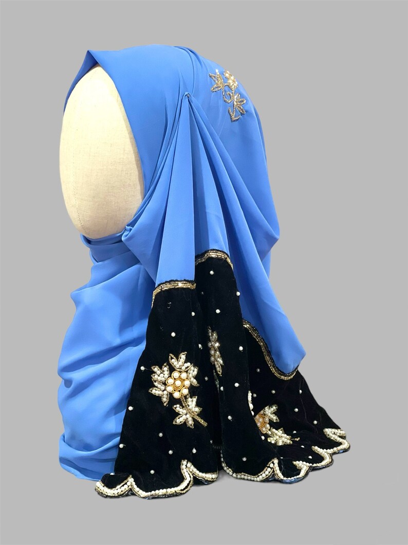 Puede incluir: Un hiyab azul claro con ribete de terciopelo negro y adornos dorados y de perlas. El hiyab tiene un borde festoneado y est&aacute; decorado con dise&ntilde;os florales.