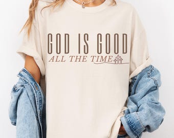 Op de afbeelding: Een beige T-shirt met de tekst "God is Good All The Time" in bruine letters. De tekst is gecentreerd op het shirt en wordt vergezeld door een kleine afbeelding van drie kruisen.