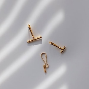 May include: Three gold-colored toggle clasps, a jewelry finding, are arranged on a white surface. The clasps have a T-bar design, with one clasp featuring a hook. The lighting creates shadows.