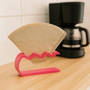 Könnte beinhalten: Ein leuchtend pinkfarbener Kaffeefilterhalter mit gewellter Oberkante, der aufrecht auf einer ebenen Fläche steht. Der Halter enthält einen einzelnen, hellbraunen, fächerförmigen Papierkaffeefilter, der sicher in der geschwungenen Form sitzt.