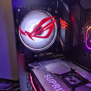 Könnte beinhalten: Ein schwarzes Computergehäuse mit einem roten und weißen Lüfter, der das ASUS ROG-Logo trägt. Der Lüfter ist an der Seite des Gehäuses montiert. Eine schwarze und silberne Grafikkarte mit dem Text "GEFORCE RTX" und "SUPRIM" ist im unteren Teil des Bildes zu sehen.