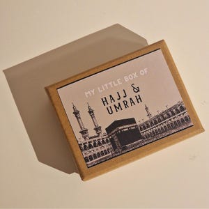 May include: A brown box with a white label that says "My Little Box of Hajj & Umrah". The label features a black and white illustration of the Kaaba and the surrounding buildings in Mecca.