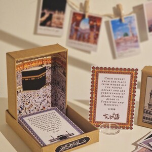 May include: A brown wooden box with a clear window showing a miniature model of the Kaaba, a cube-shaped building in Mecca, Saudi Arabia. The box is open and there are two cards inside with text from the Quran. The text on the cards reads: "And remember when Abraham raised the foundation of the house with Ishmael. Both praying, our Lord! Accept this from us. You are indeed the All-Hearing, All-Knowing. 2:127" and "Then depart from the place from where all the people depart and ask forgiveness of Allah. Indeed, Allah is forgiving and merciful. 2:199".