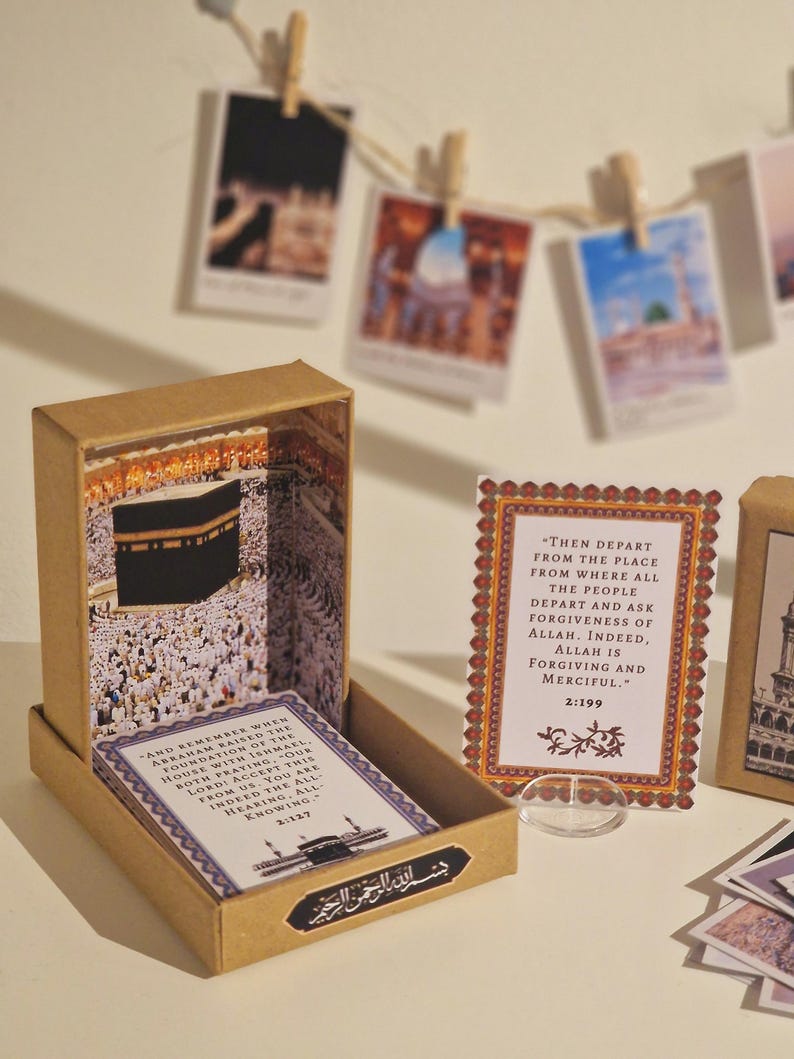 May include: A brown wooden box with a clear window showing a miniature model of the Kaaba, a cube-shaped building in Mecca, Saudi Arabia. The box is open and there are two cards inside with text from the Quran. The text on the cards reads: "And remember when Abraham raised the foundation of the house with Ishmael. Both praying, our Lord! Accept this from us. You are indeed the All-Hearing, All-Knowing. 2:127" and "Then depart from the place from where all the people depart and ask forgiveness of Allah. Indeed, Allah is forgiving and merciful. 2:199".