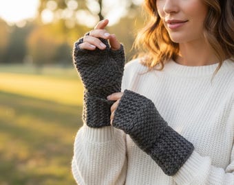 May include: Dark gray fingerless gloves made of a chunky knit material. The gloves are worn by a person wearing a white sweater. The background is blurred, with hints of green and yellow, suggesting an outdoor setting.
