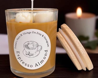 May include: Espresso Almond scented candle in a clear glass jar. The candle has a light brown wax with two white wax cubes on top. A round label with a coffee cup illustration is on the jar. A wooden match holder is next to the candle.