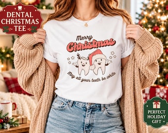 May include: A white t-shirt featuring a graphic of two cartoon teeth, one wearing reindeer antlers and the other a Santa hat. The text reads Merry Christmas and May all your teeth be white, surrounded by small star icons in red and brown.