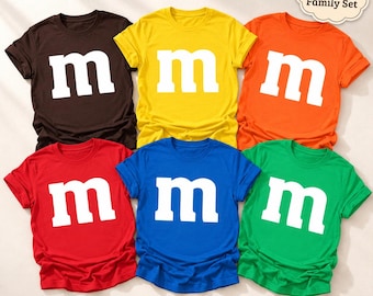 May include: A set of six short-sleeved crew neck t-shirts in brown, yellow, orange, red, blue, and green. Each shirt features a large white lowercase letter m printed on the front. A small cloud-shaped tag in the corner reads Matching Family Set.