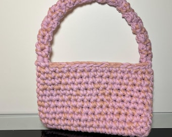 May include: A handmade, rectangular crochet bag in a soft pink hue with a braided handle. The bag's body features a tight stitch pattern, and the handle is a blend of pink and beige. The bag is designed for carrying items.