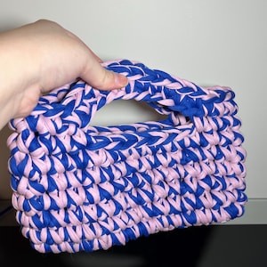 May include: A handmade, rectangular handbag in a woven pattern. The bag features alternating rows of blue and pink fabric, with a matching blue and pink handle. The bag is held by a hand.