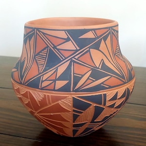 May include: A terracotta-coloured clay pot featuring a wide, rounded body and a shorter neck. The surface is decorated with intricate, sharp-edged geometric patterns in matte black and natural clay tones, including triangles and linear hatch marks.