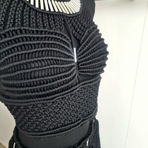 以下が含まれることがあります： 黒いマクラメのクロップトップとスカートのセット、シルバーの金属アクセント付き。ホルターネックのトップスは、シルバーの襟が付いた幾何学模様です。スカートはフリンジのディテールとシルバーの装飾が施されています。セットは黒いマネキンに展示されています。