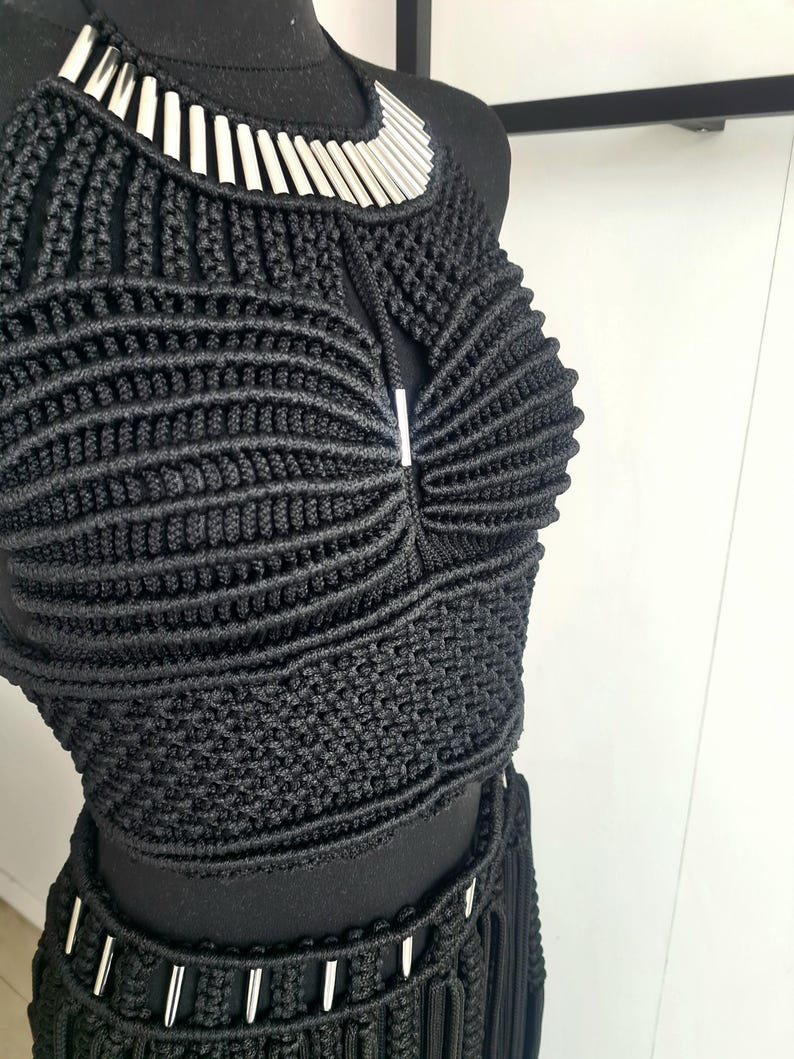 以下が含まれることがあります： 黒いマクラメのクロップトップとスカートのセット、シルバーの金属アクセント付き。ホルターネックのトップスは、シルバーの襟が付いた幾何学模様です。スカートはフリンジのディテールとシルバーの装飾が施されています。セットは黒いマネキンに展示されています。