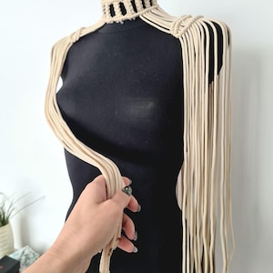 May include: A beige macrame choker necklace with long fringe strands extending from the sides. The necklace is displayed on a black mannequin.