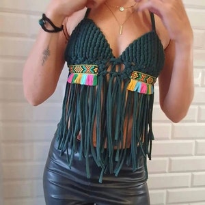 以下が含まれることがあります： フリンジのディテールが施されたダークグリーンのクロシェトップ。トップにはカラフルな刺繍のバンドとタッセルが付いています。