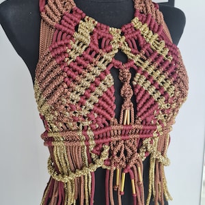May include: A maroon and gold macrame top with fringe detailing. The top is made with a variety of cords and has a geometric design.