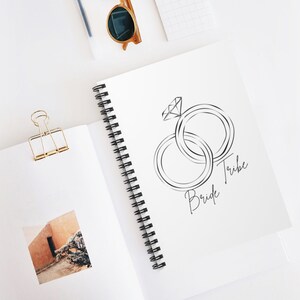 May include: A white spiral bound notebook with a black line drawing of two interlocking rings with a diamond on one ring. The text "Tribe Bride" is written in cursive below the rings.