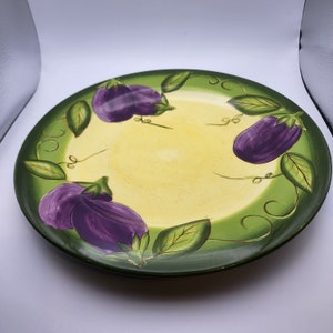 May include: A round ceramic plate with a green rim and a yellow center. The plate is decorated with purple eggplants and green leaves.