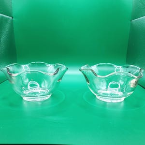 May include: Two clear glass bowls with a ruffled edge. The bowls are round with a scalloped rim and sit on a green surface. The bowls are empty and appear to be made of crystal or a similar material.