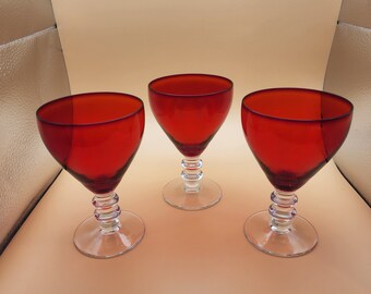 May include: Three red wine glasses with clear stems and bases. The glasses have a rounded bowl and a thick, clear rim.