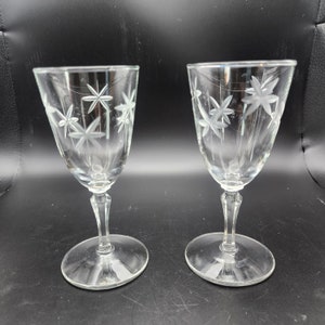 May include: Two clear glass wine glasses with etched star designs. The glasses have a tall, slender stem and a round base.