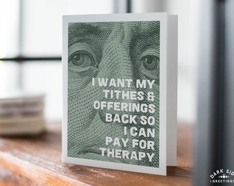 May include: A white greeting card with the text "I WANT MY TITHES & OFFERINGS BACK SO I CAN PAY FOR THERAPY" over a green image of a dollar bill. The card is on a wooden surface.