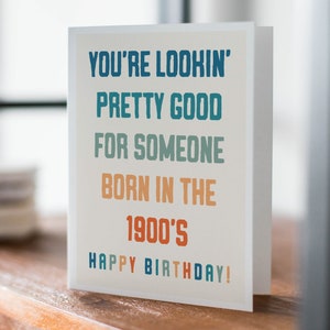 May include: A folded birthday card with the text "YOU'RE LOOKIN' PRETTY GOOD FOR SOMEONE BORN IN THE 1900'S HAPPY BIRTHDAY!" in blue, green, and orange lettering. The card is on a wooden surface.