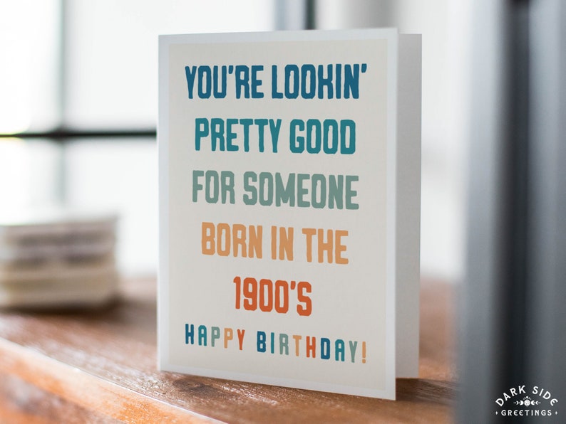 May include: A folded birthday card with the text "YOU'RE LOOKIN' PRETTY GOOD FOR SOMEONE BORN IN THE 1900'S HAPPY BIRTHDAY!" in blue, green, and orange lettering. The card is on a wooden surface.