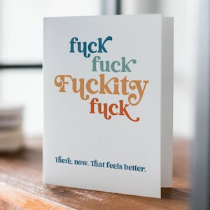 May include: A white greeting card with the words "fuck, fuck, Fuckity, fuck" in blue, teal, orange, and red lettering. The phrase "There, now. That feels better." is printed below in blue. The card is on a wooden surface.