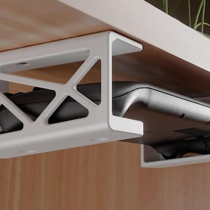 May include: A white under-desk mount with a geometric, open-frame design, securely holding a black handheld gaming console. The mount is attached to the underside of a light-colored wooden surface, providing a storage solution for the device.