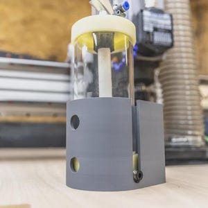 May include: A gray and clear cylindrical device with a yellow top and a clear container. The device has two circular holes and a black fitting. The background shows a woodworking machine.