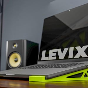 May include: A laptop computer rests on a bright green laptop stand. The stand is angled for ergonomic use. The laptop screen displays the text "LEVIX XL" in white and green. A black speaker with a yellow speaker cone is in the background.