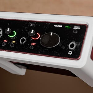 May include: A white Scarlett Solo audio interface mounted under a desk. The device features a black front panel with a large monitor knob, input ports, and indicator lights labeled 48V, AIR, INST, and Direct Monitor, housed in a custom white frame.