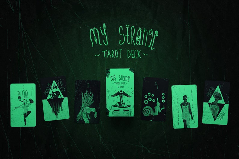 May include: A set of tarot cards with black and white illustrations on a green background. The cards are arranged in a row with the box in the middle. The box has the text "My Strange Tarot Deck" on it. The cards are labeled with numbers and names, such as "The Fool" and "Knight of Swords".