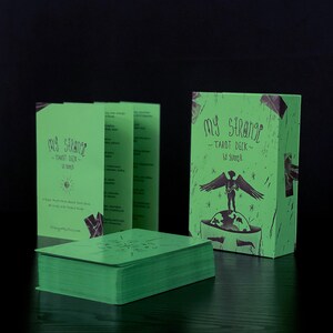 May include: A green box with the text "My Strange Tarot Deck" and a black and white illustration of a person with wings standing on a globe. The box is sitting on a dark wooden surface.