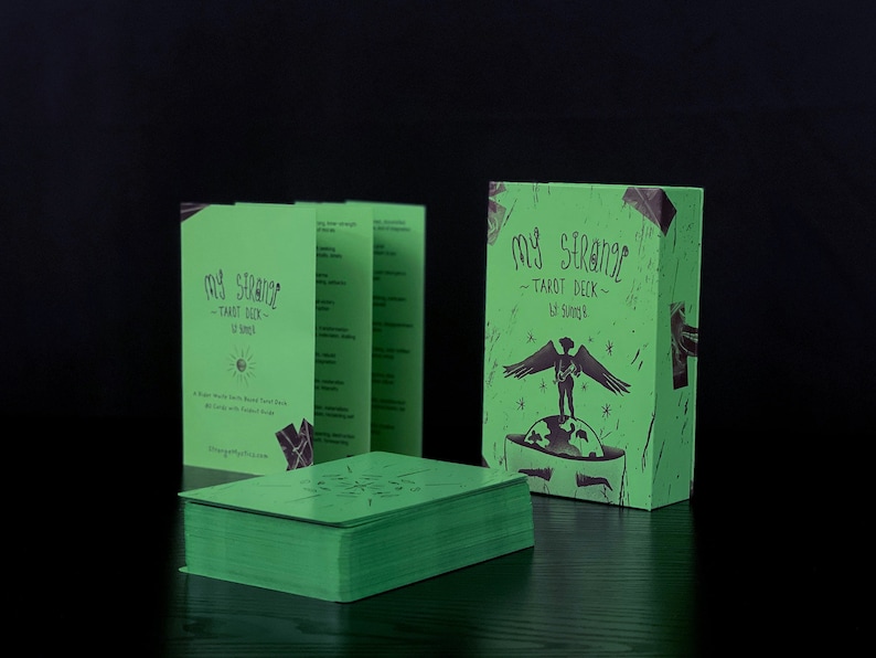 May include: A green box with the text "My Strange Tarot Deck" and a black and white illustration of a person with wings standing on a globe. The box is sitting on a dark wooden surface.