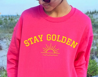 May include: A pink sweatshirt with the text "STAY GOLDEN" in yellow lettering and a yellow graphic of a sun rising over water.