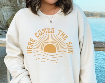 Könnte beinhalten: Ein cremefarbener Pullover mit einer Grafik einer gelben Sonne, die über Wellen aufgeht, und dem Text "Here Comes The Sun".