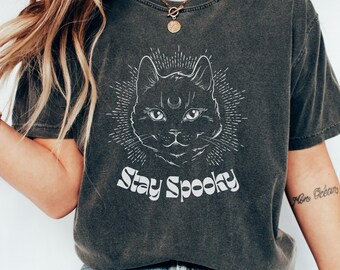 Könnte beinhalten: Ein dunkelgraues T-Shirt mit einem weißen Grafikdruck eines schwarzen Katers mit einer Mondsichel auf der Stirn. Der Text "Stay Spooky" ist unter dem Katzenmotiv gedruckt.