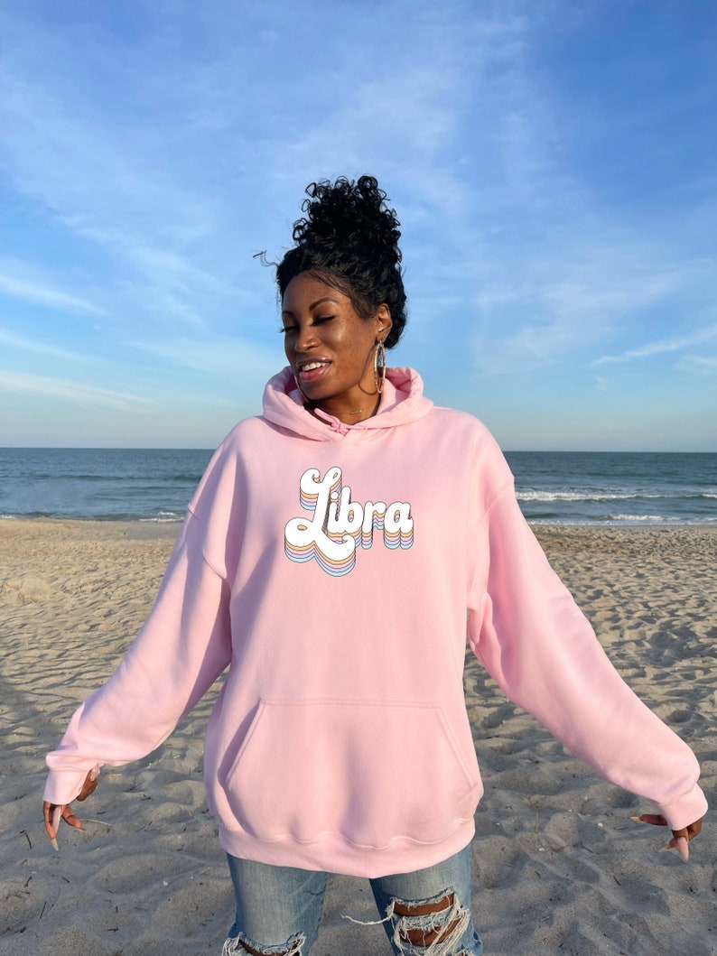 K&ouml;nnte beinhalten: Ein rosa Hoodie mit einem Retro-Style "Libra"-Motiv in Regenbogenfarben. Der Hoodie wird von einer Person getragen, die auf einem Sandstrand steht.