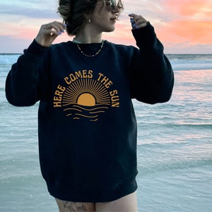 Puede incluir: Sudadera negra con un gr&aacute;fico amarillo de un sol saliendo sobre el oc&eacute;ano y el texto "Here Comes The Sun".