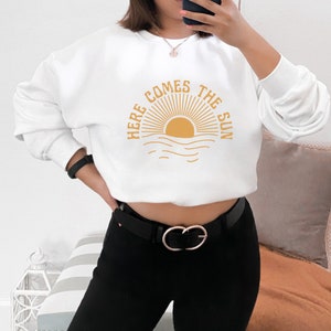 Puede incluir: Sudadera blanca con un gr&aacute;fico amarillo de un sol saliendo sobre el oc&eacute;ano y el texto "Here Comes The Sun".