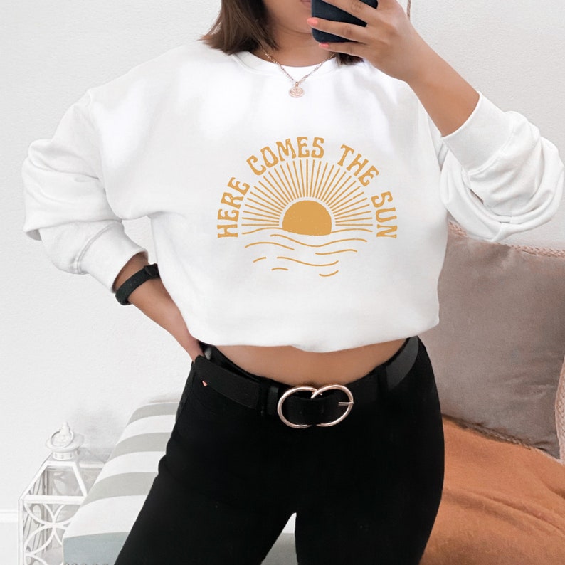 Puede incluir: Sudadera blanca con un gr&aacute;fico amarillo de un sol saliendo sobre el oc&eacute;ano y el texto "Here Comes The Sun".