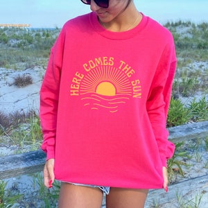Puede incluir: Una sudadera rosa con un gr&aacute;fico amarillo de un sol saliendo sobre el oc&eacute;ano. El gr&aacute;fico dice "Here Comes The Sun".