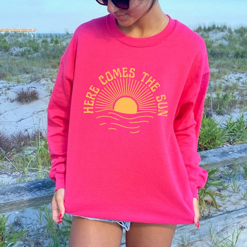 Puede incluir: Una sudadera rosa con un gr&aacute;fico amarillo de un sol saliendo sobre el oc&eacute;ano. El gr&aacute;fico dice "Here Comes The Sun".