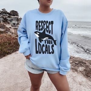May include: Light blue sweatshirt with a black graphic of an orca and the text "Respect the Locals".